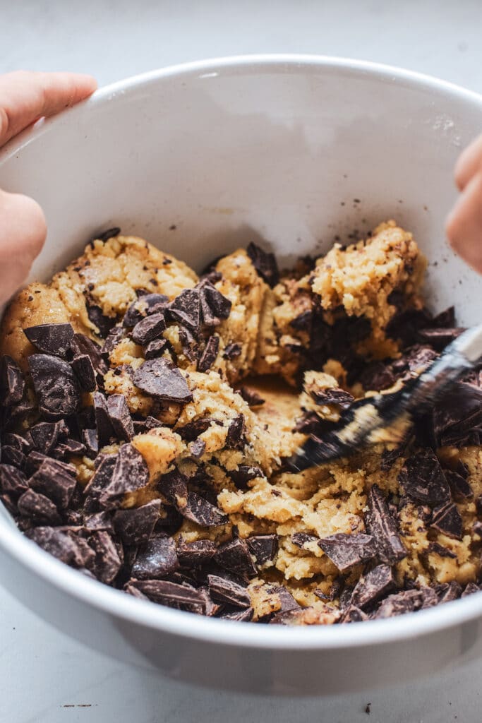 Chocolate chunks being folded into a white bowl of cookie dough