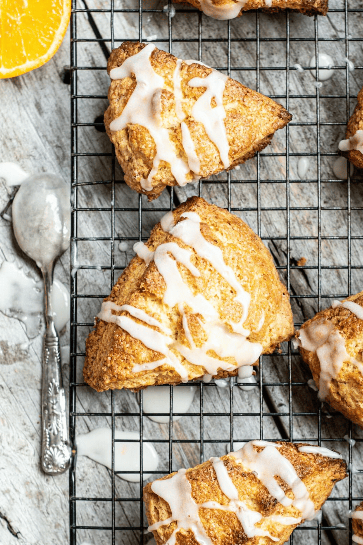 Triangular homemade scones drizzled with white icing cool on a wire rack, with a vintage spoon coated in glaze and a sliced orange nearby on a rustic surface.