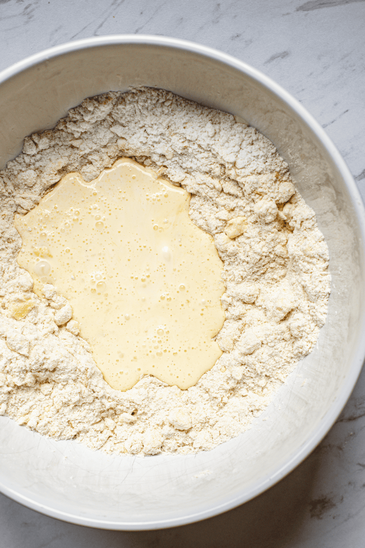 A white mixing bowl containing flour with a well in the center filled with a liquid mixture, likely eggs and milk, perfect for making homemade scones, sits on a light-colored countertop.