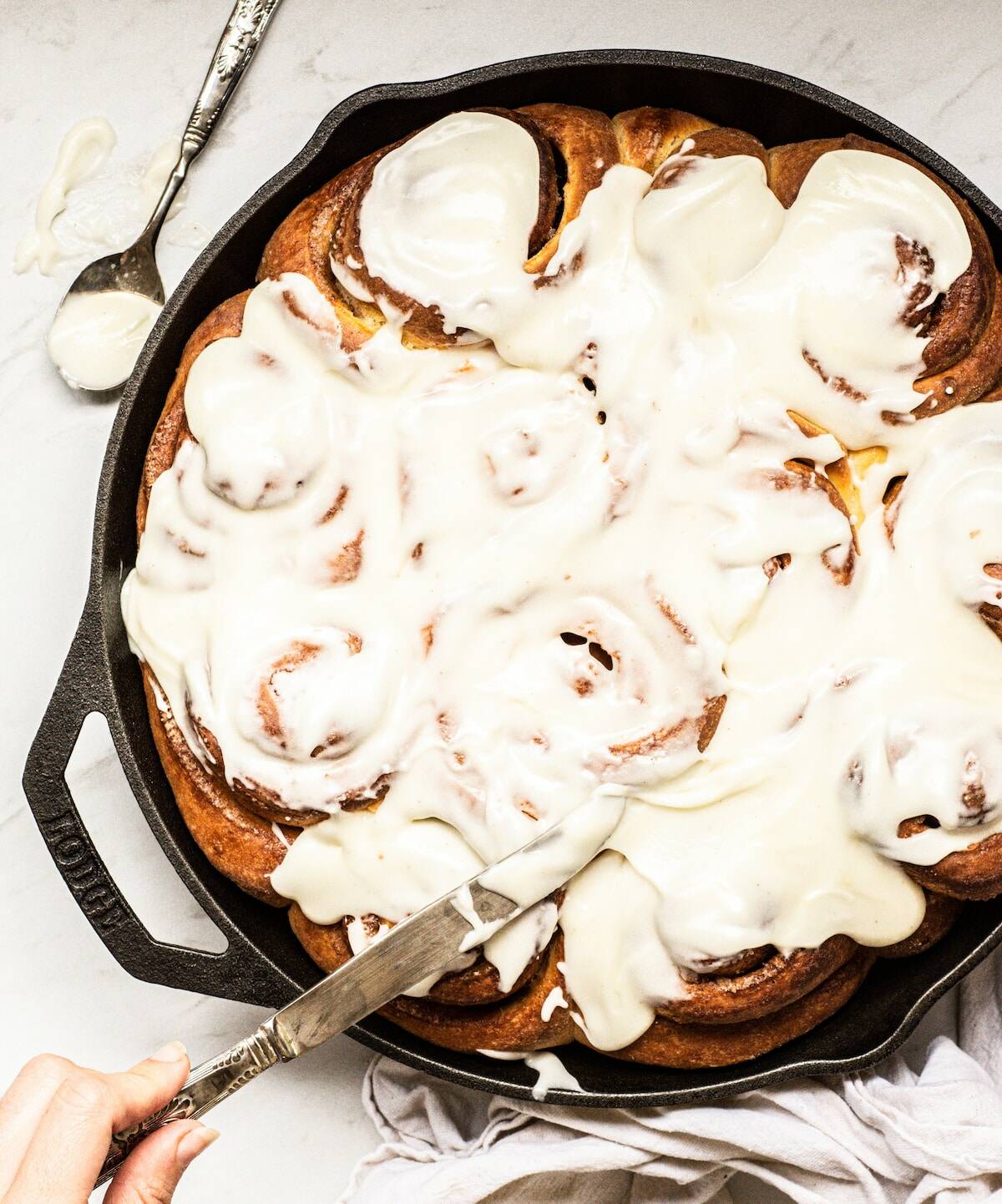 a cast iron pan with baked cinnamon rolls with white icing. A hand is holding a knife and adding on the icing