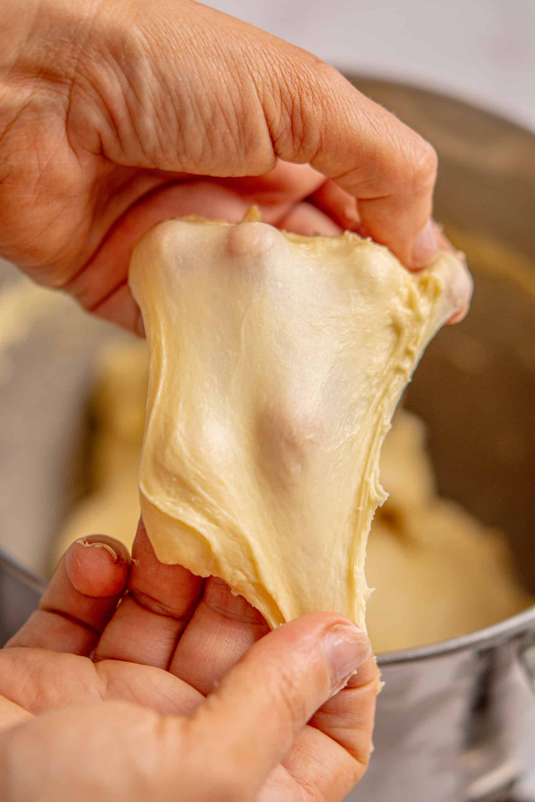 Close-up of hands stretching a piece of dough to show its elasticity and smooth texture, demonstrating the gluten windowpane test above a mixing bowl when making a brioche bread recipe.
