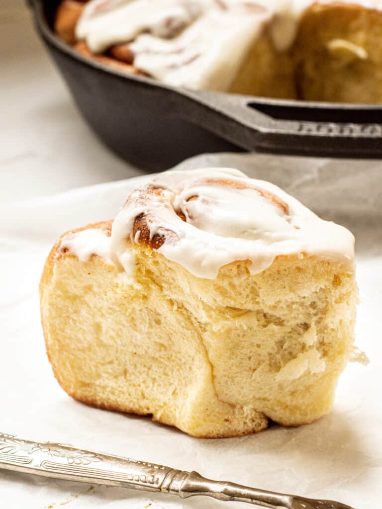 a close up of a baked brioche cinnamon roll with a fork in the foreground and a cast iron pan in the background