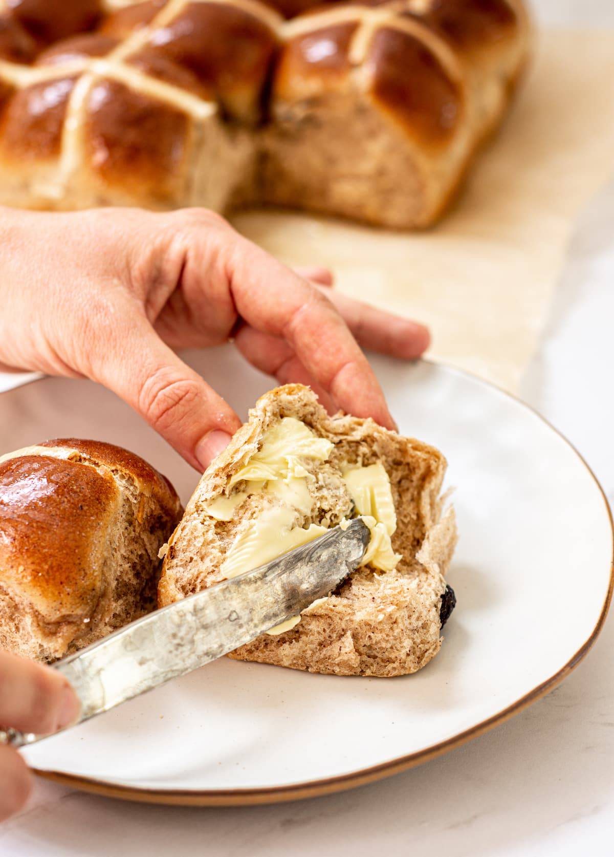 a hand holding a knife, smearing butter over a cut hot cross bun