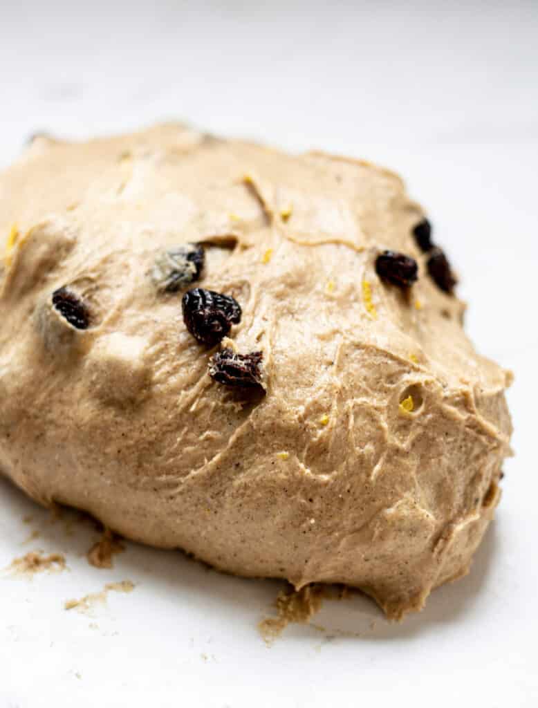 a mound of kneaded hot cross bun dough with raisins on a white bench