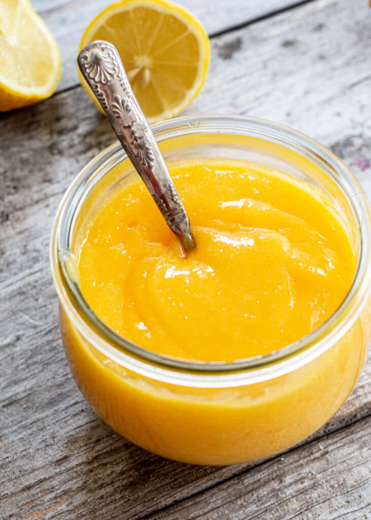 a close up look of a jar of lemon curd with a silver spoon in the middle. On a wooden board with two lemons in the background