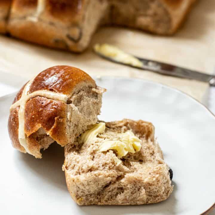 a plate with a buttered hot cross bun, other buns are in the background.