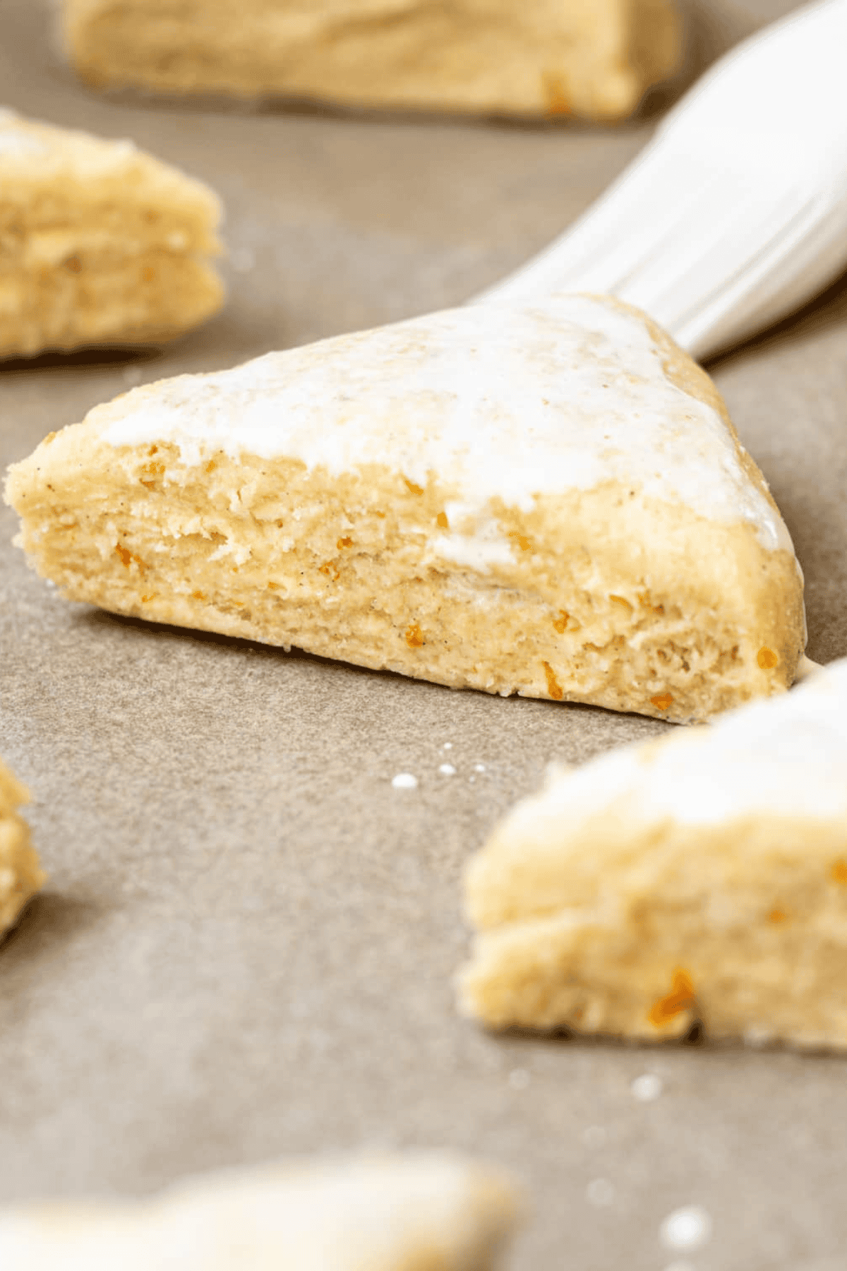 A close-up of a triangular glazed homemade scone with light orange zest flecks, resting on parchment paper, with other scones blurred in the background.