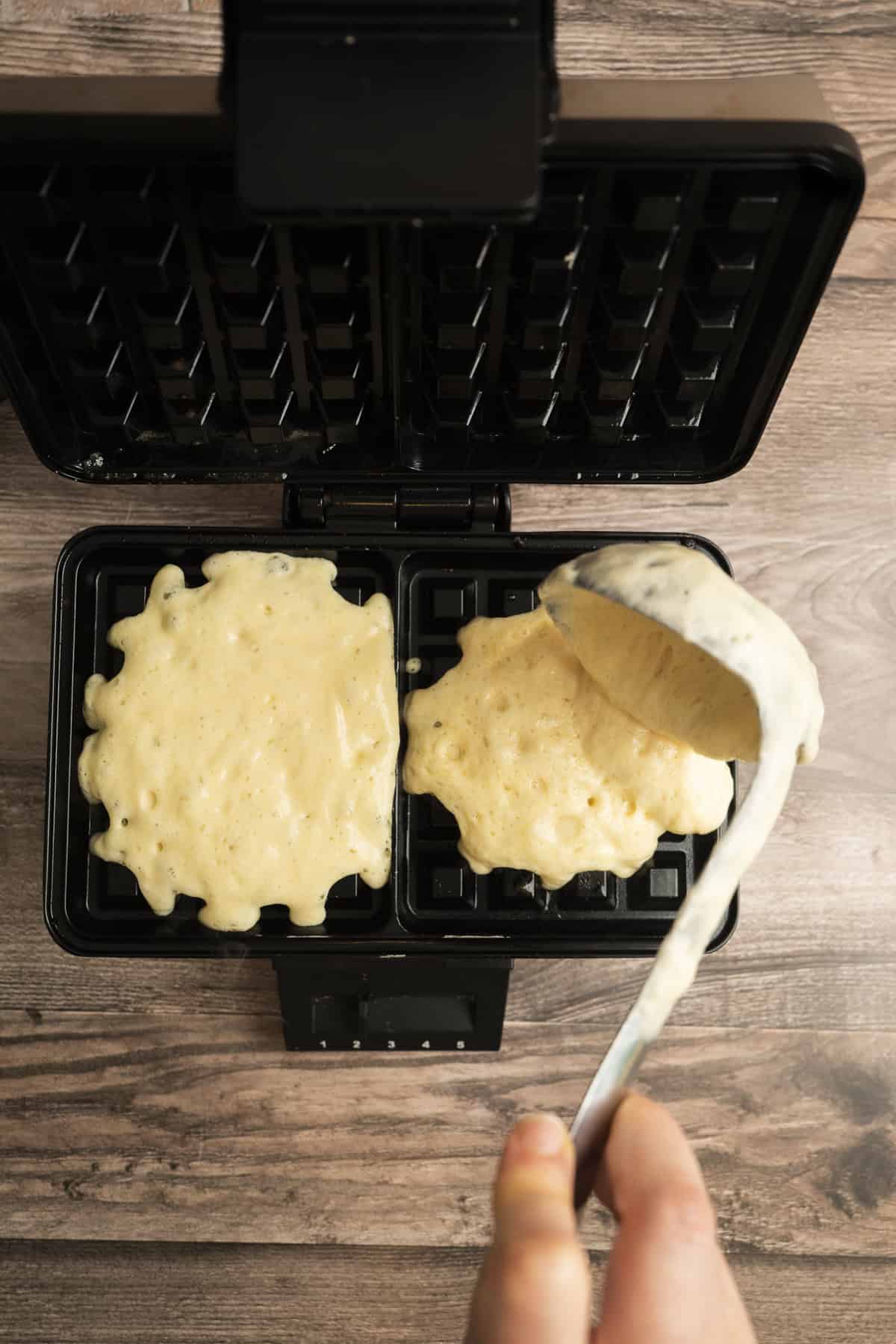 A hand pours yeasted Belgian waffle batter onto a preheated waffle iron with two sections, one already covered in batter. The waffle iron sits on a wooden surface.