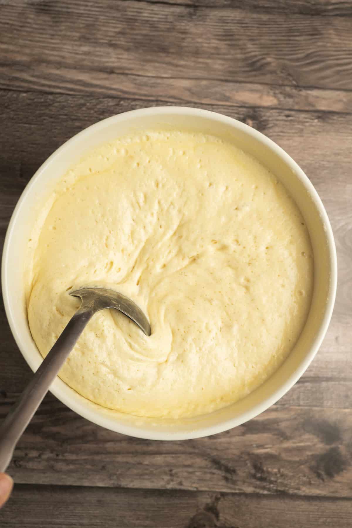 A metal spoon stirs thick, yellow Yeasted Belgian Waffles batter in a white bowl on a wooden surface.