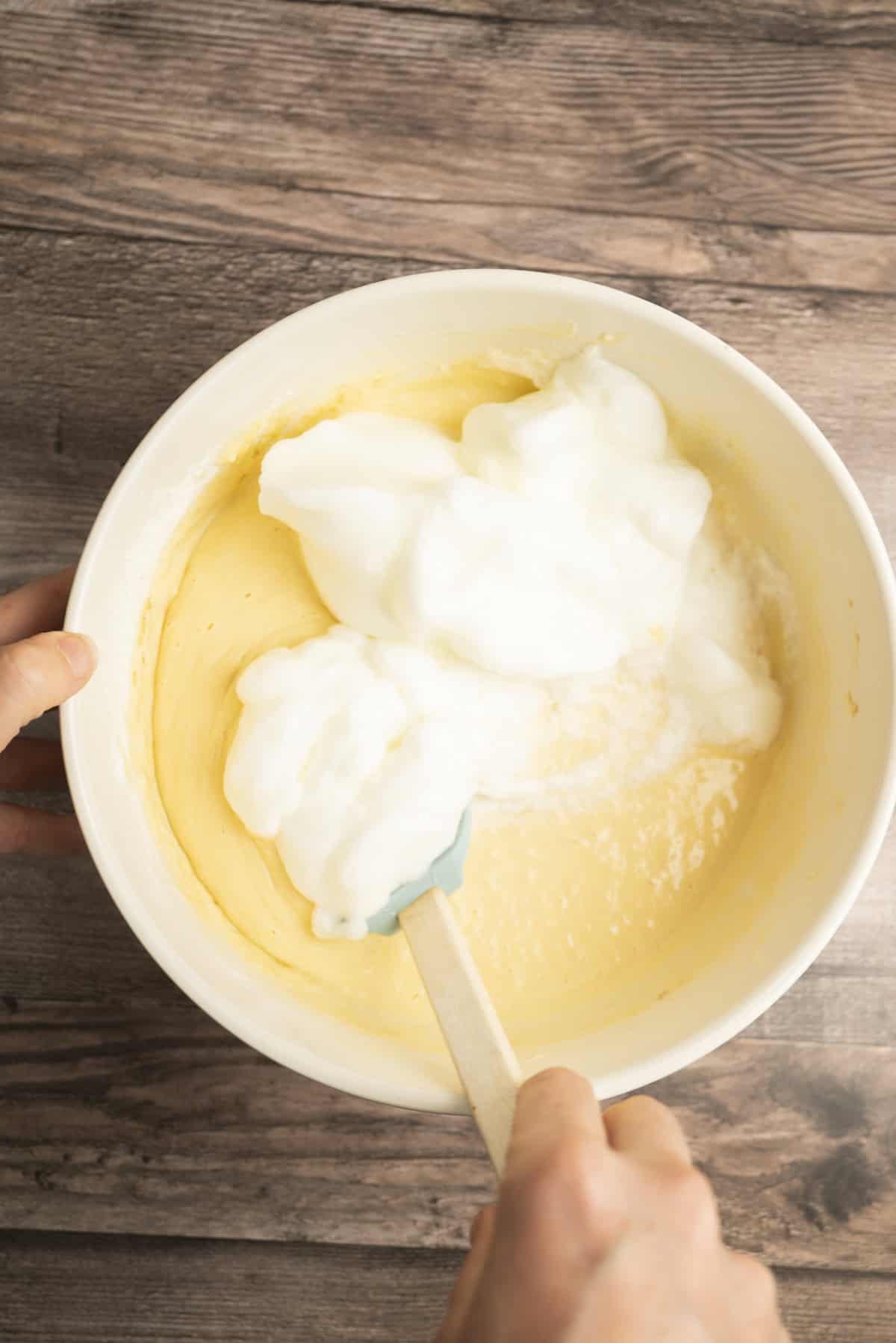 A person uses a spatula to fold fluffy egg whites into a bowl of yellow cake batter, just as you would when preparing classic yeasted Belgian waffles, on a wooden surface.