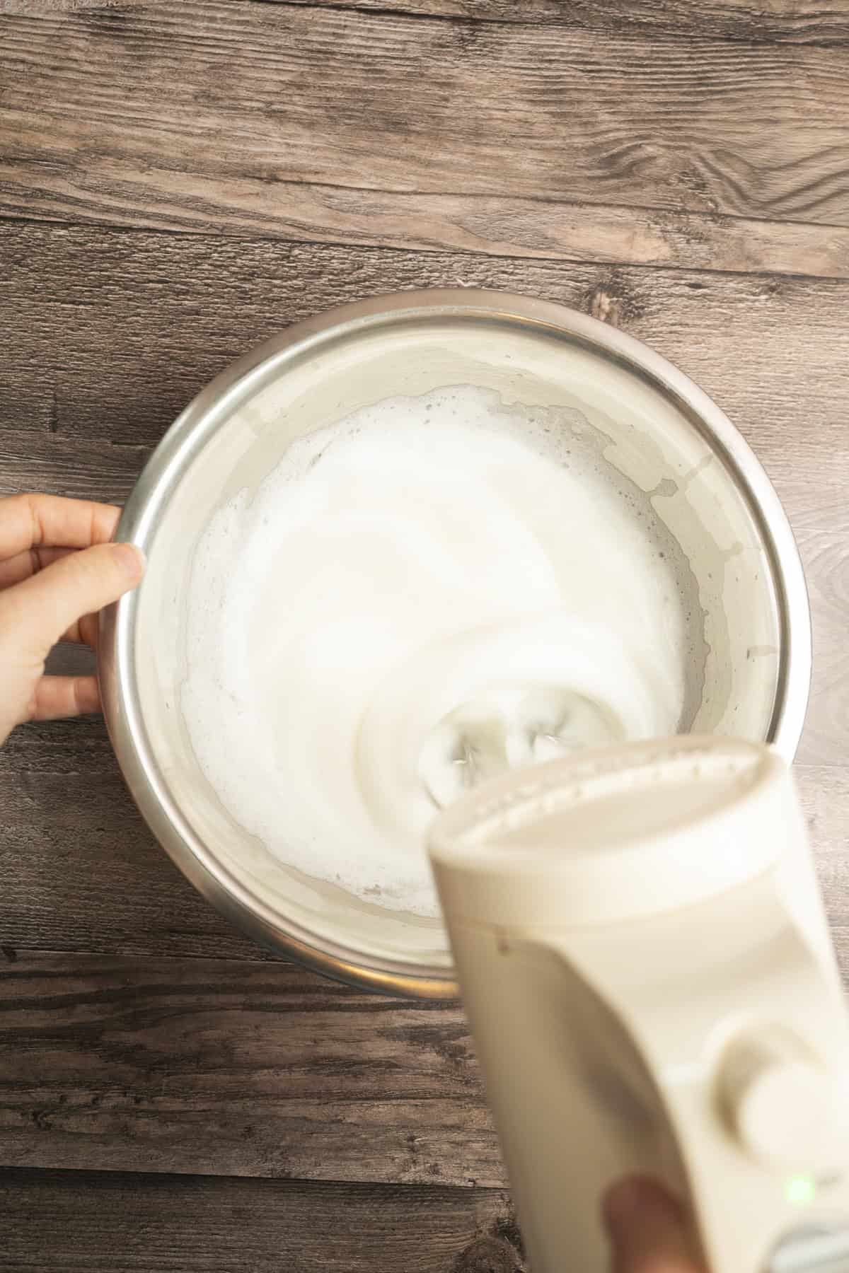 A person uses a handheld electric mixer to beat egg whites in a metal bowl on a wooden surface. The foamy, partially whipped egg whites are perfect for making fluffy Yeasted Belgian Waffles.