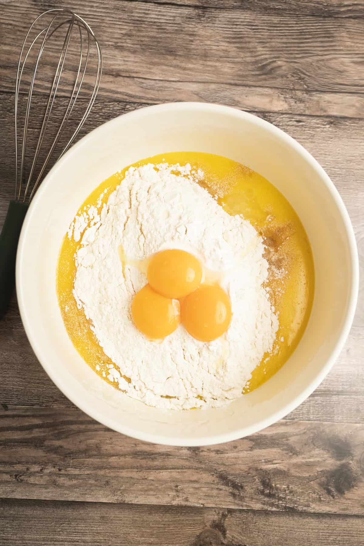 A white mixing bowl contains flour, three egg yolks, melted butter, and ingredients for yeasted Belgian waffles, with a metal whisk resting beside the bowl on a wooden surface.