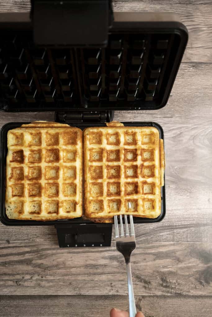A close-up of two golden Yeasted Belgian Waffles in a waffle maker on a wooden surface. A hand holds a fork, ready to lift one waffle from the waffle maker.