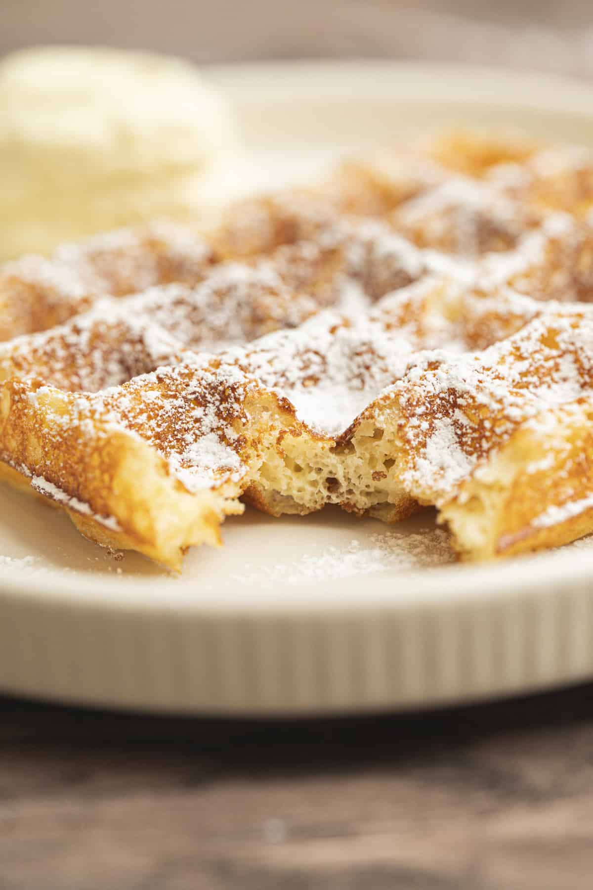 A close-up of a golden Yeasted Belgian Waffle dusted with powdered sugar, with a bite taken out of it, served on a cream-colored plate. In the background, there is a scoop of vanilla ice cream.