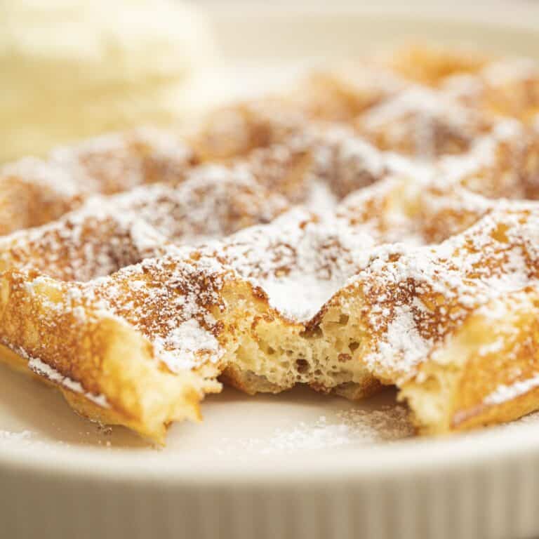 A close-up of a golden, fluffy Yeasted Belgian Waffle dusted with powdered sugar, with a bite taken out of the middle, on a white plate.