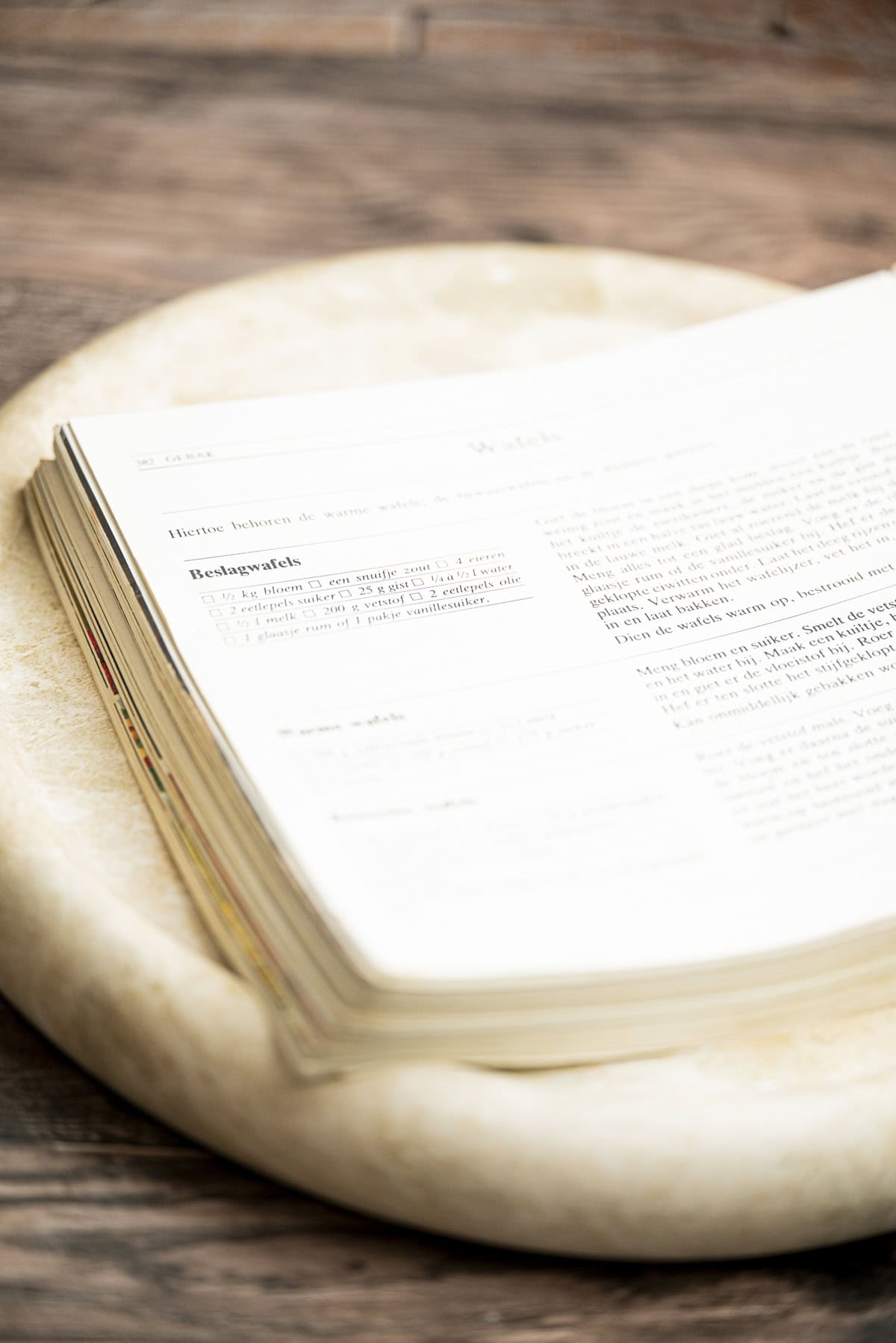 An open book with text in flemish lies on a round, light-colored stone surface atop a wooden table. The page edges are slightly curled, and the focus is on the bottom left corner.
