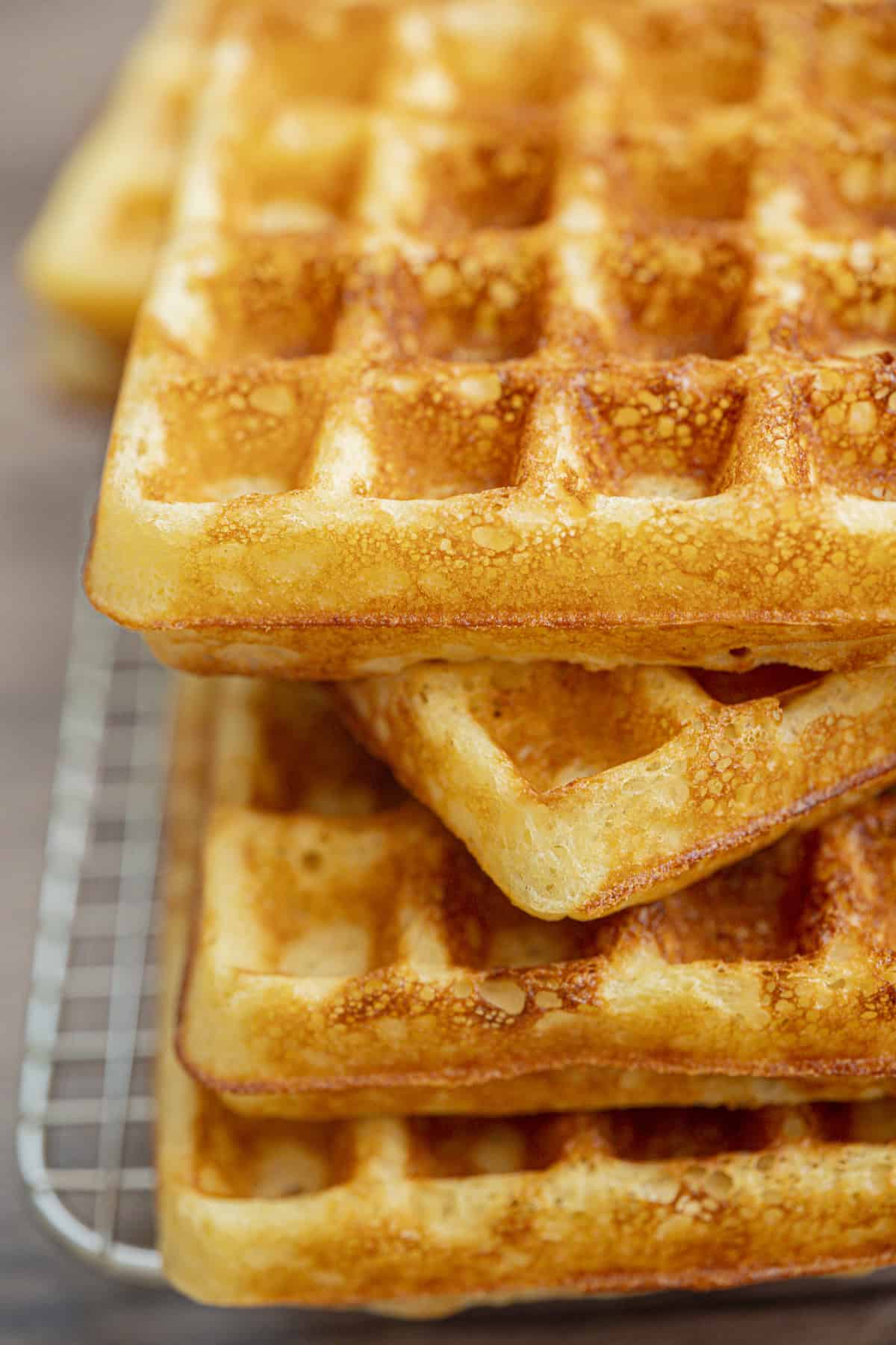 A close-up of golden brown Yeasted Belgian Waffles stacked on a cooling rack, showing their crispy texture and deep square indentations.