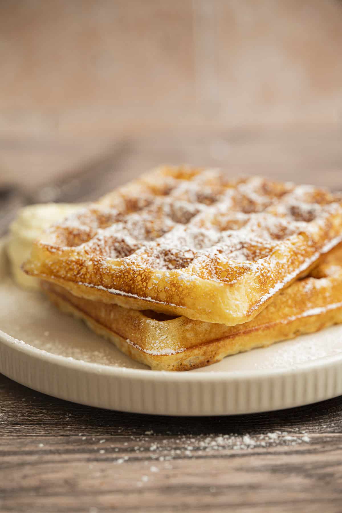Two golden, crispy Yeasted Belgian Waffles dusted with powdered sugar are stacked on a white plate, sitting on a wooden surface. The background is softly blurred.