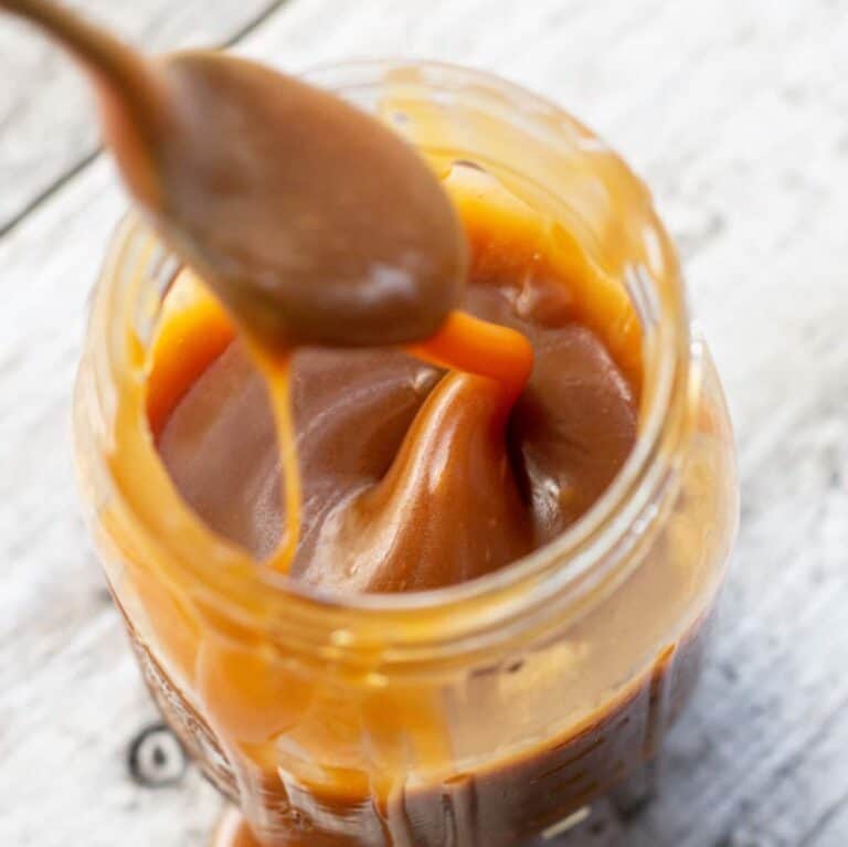 A close-up of a jar filled with thick, creamy 3 Ingredient Caramel Sauce. A spoon is dipped into the jar, lifting some caramel that drips back inside. The jar sits on a light wooden surface.