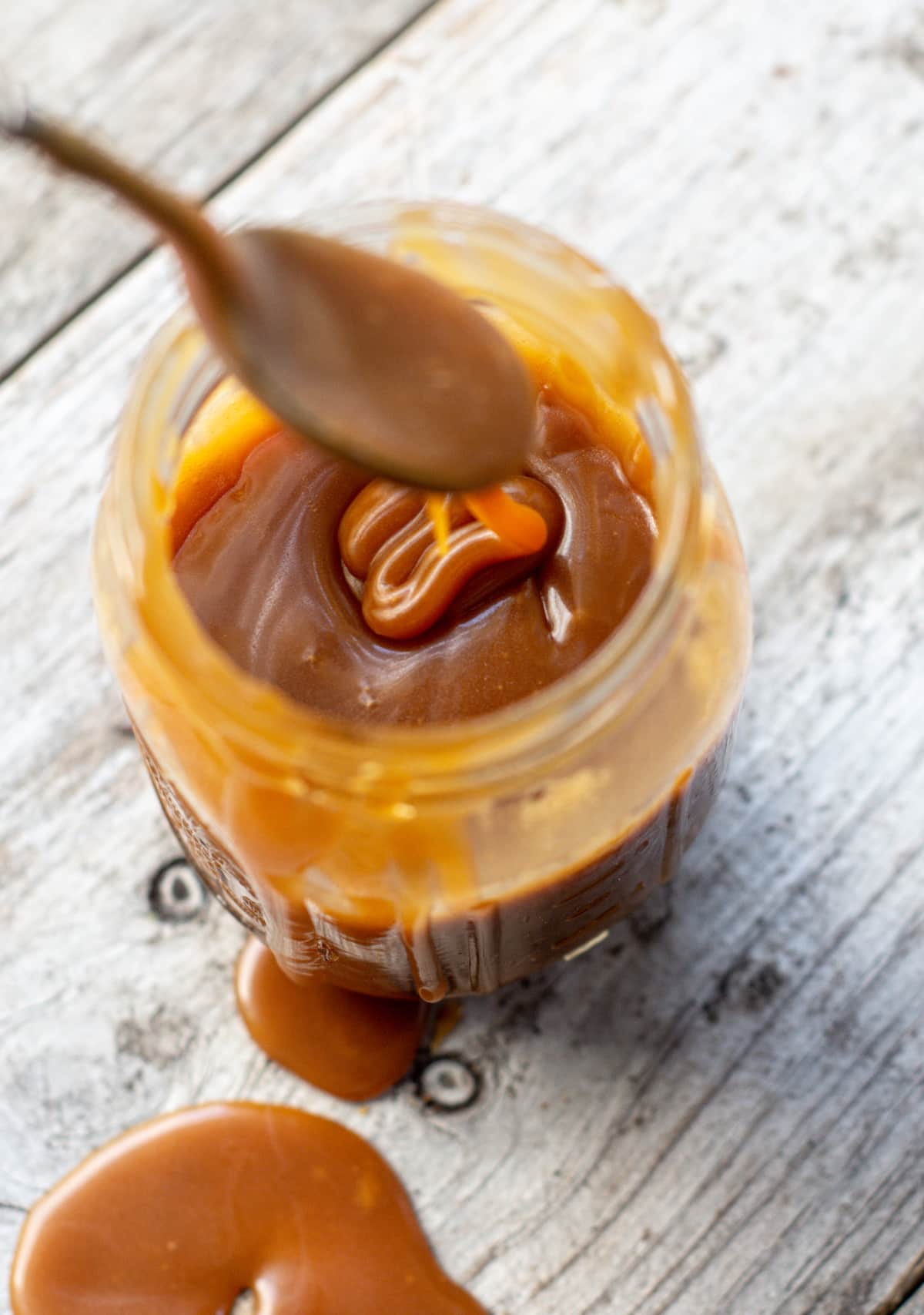 A jar of thick caramel sauce sits on a wooden surface, with caramel dripping down the side and pooling on the table. A spoon coated in caramel hovers above the jar.