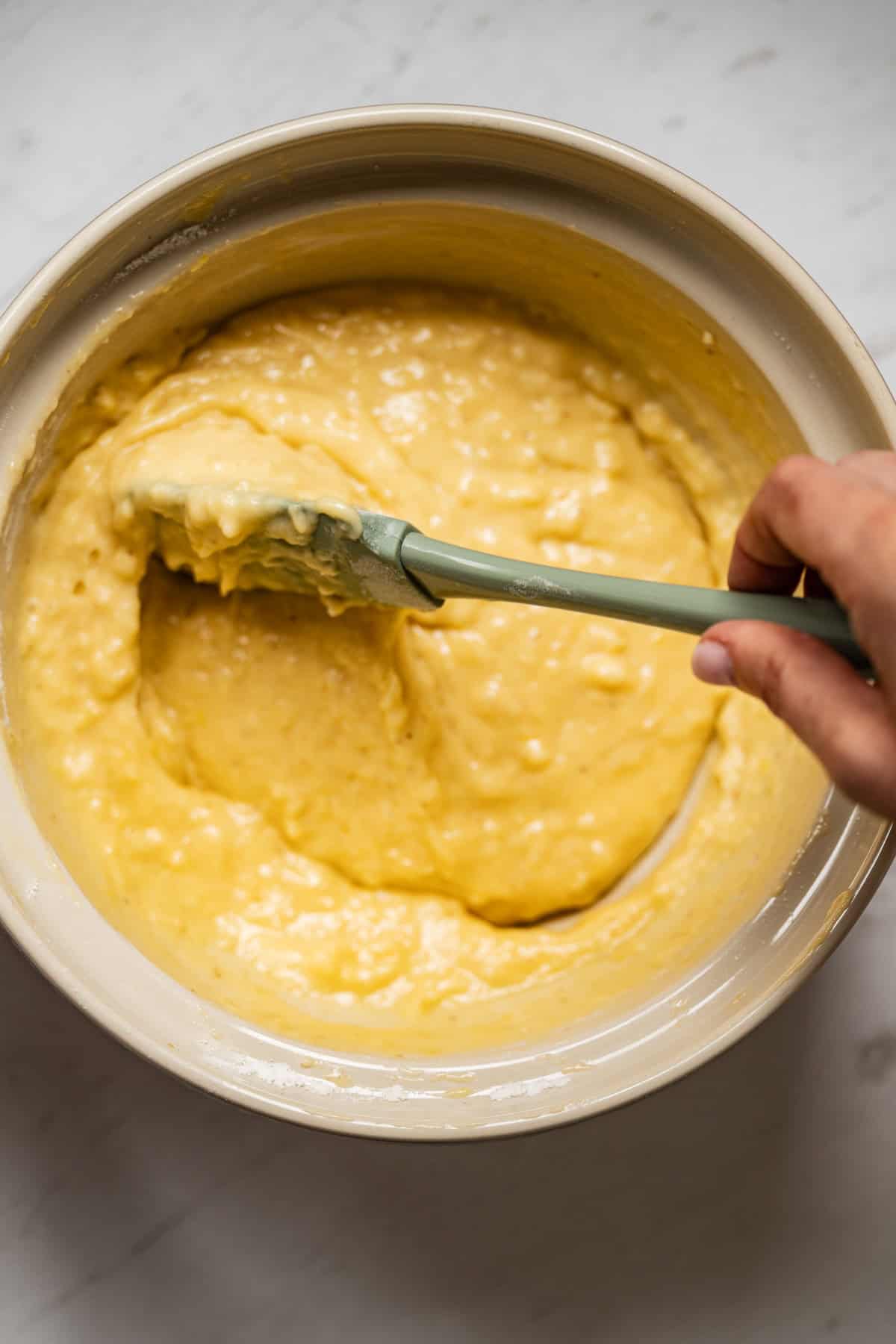 A hand uses a green spatula to mix thick, yellow lemon yogurt olive oil cake batter in a large white bowl on a light-colored surface.