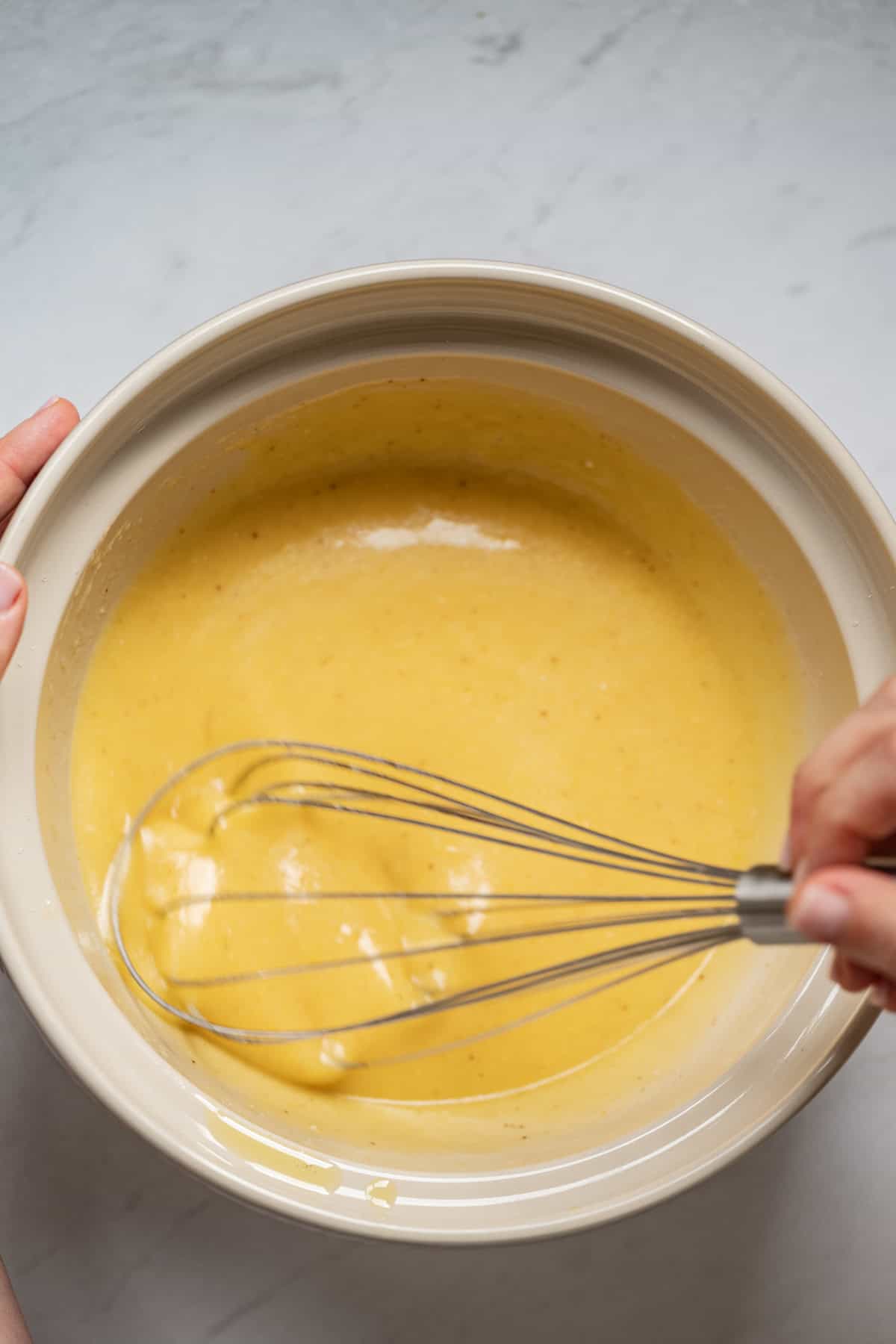 A person whisking a smooth, yellow batter for a lemon yogurt olive oil cake in a large, light-colored mixing bowl on a light countertop.