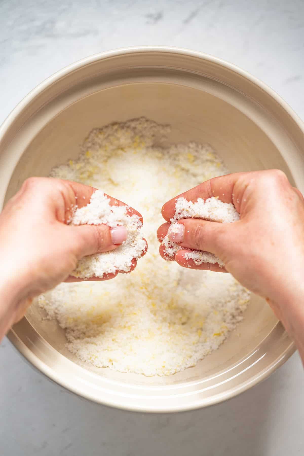 Two hands are rubbing together a crumbly mixture of flour and butter over a large beige mixing bowl, likely preparing dough or pastry for a lemon yogurt olive oil cake on a light surface.