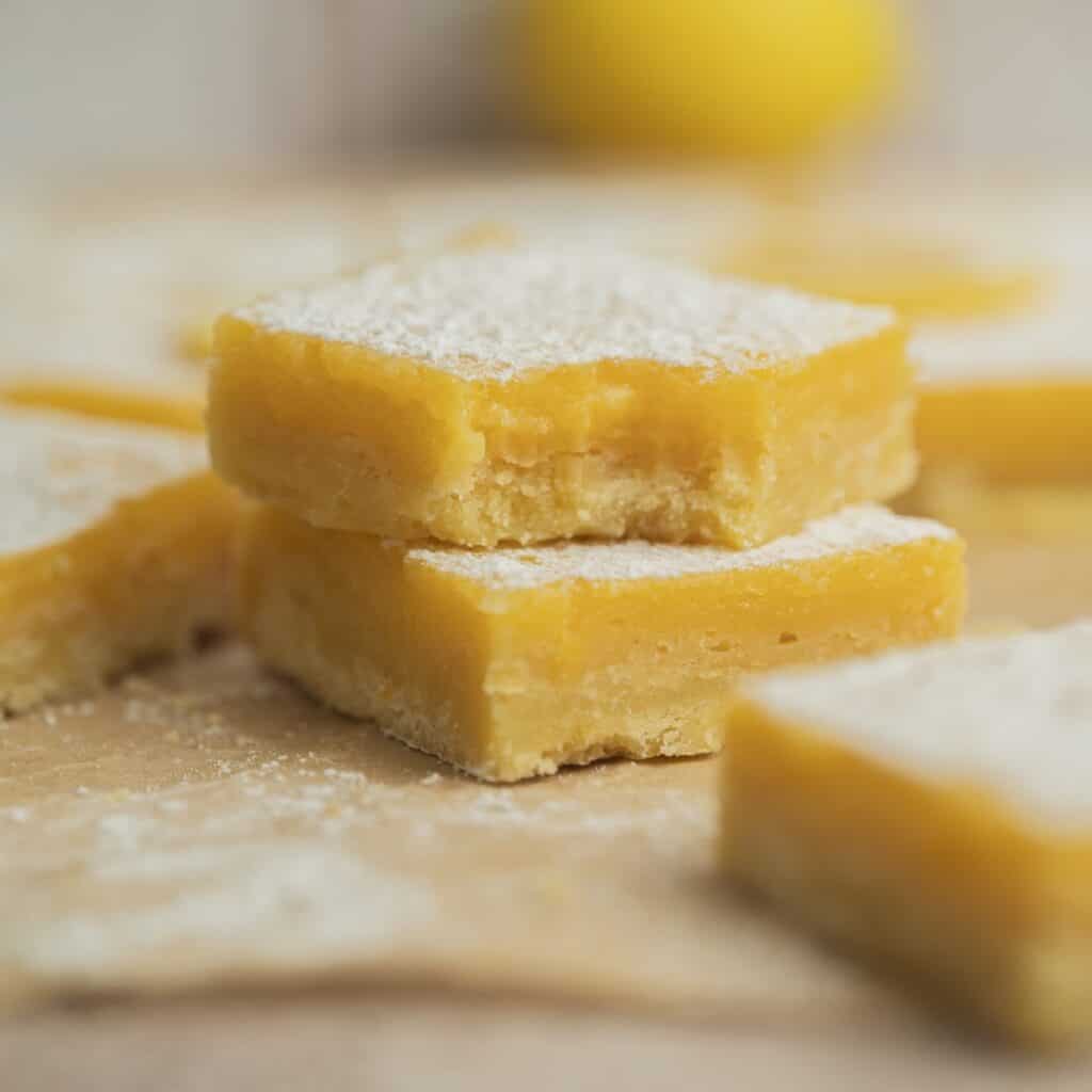 Three easy Meyer lemon bars stacked, topped with powdered sugar, with more lemon bars and a blurred lemon in the background. The bars have a yellow, creamy layer on a thick, crumbly crust.