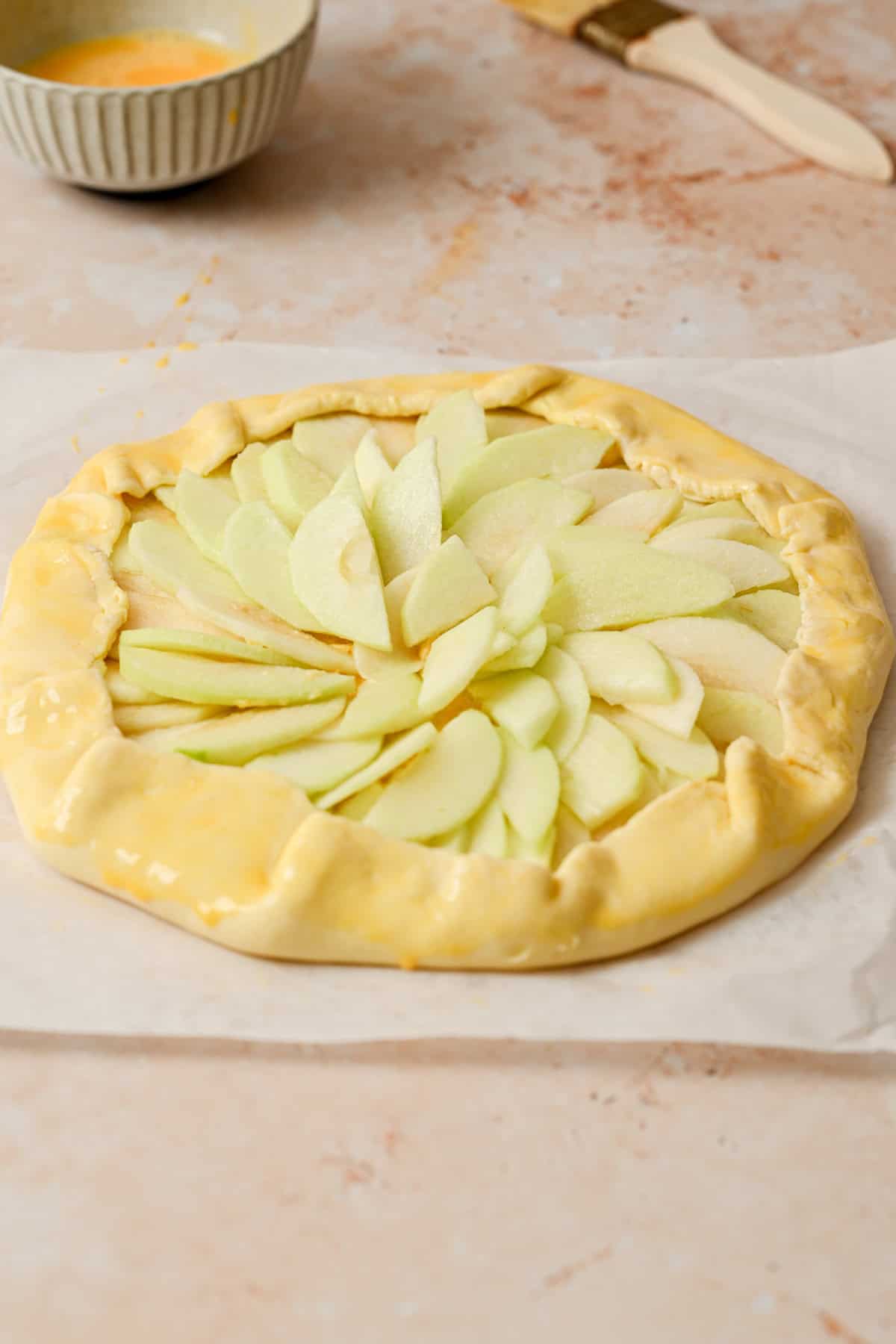 Unbaked apple galette with almond filling, featuring thinly sliced apples arranged in a spiral on a rustic pastry crust, edges folded over. A bowl and pastry brush are visible in the background on a light marble surface.