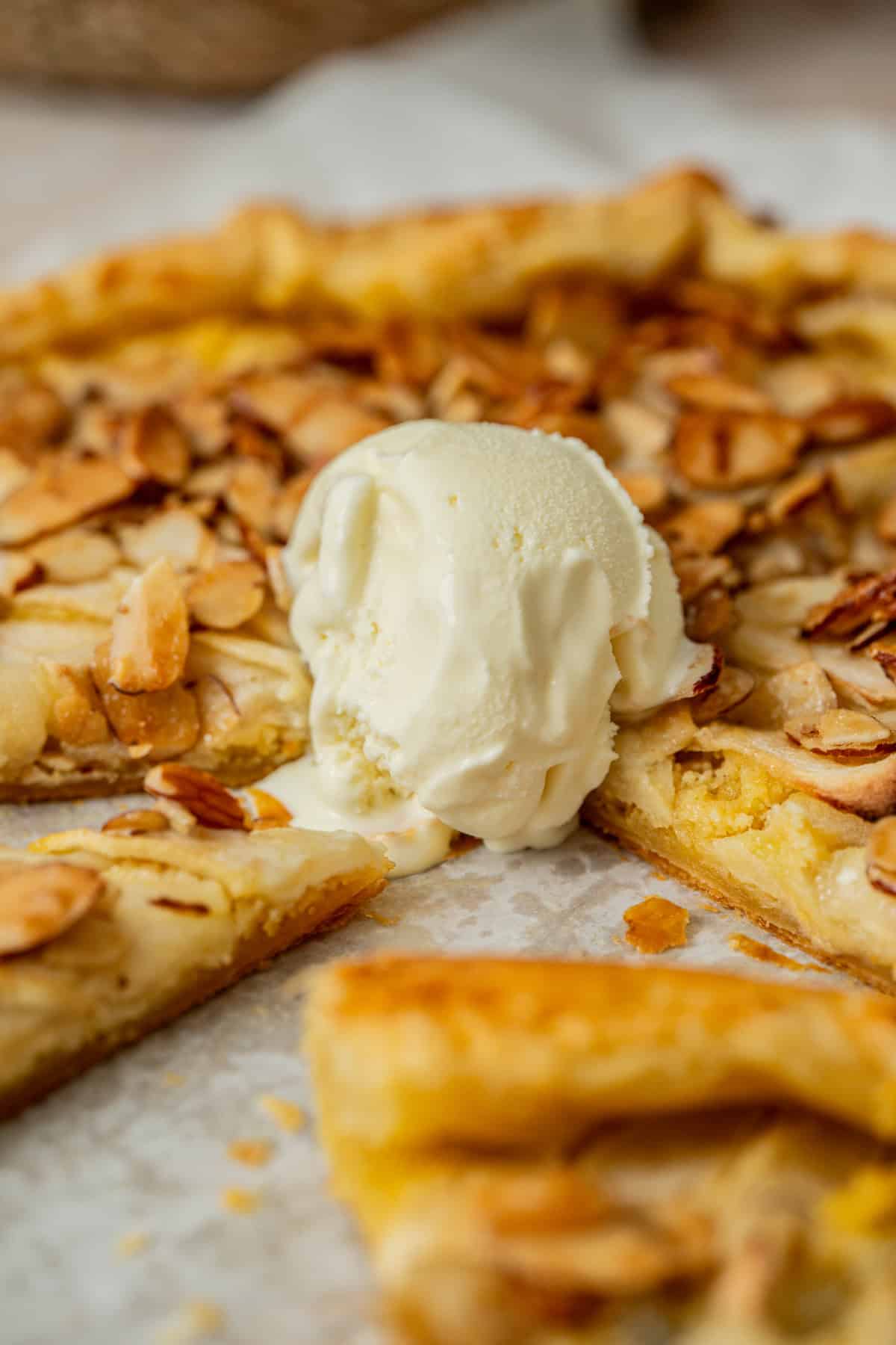 Close-up of a sliced almond tart, reminiscent of an apple galette with almond filling, topped with a scoop of melting vanilla ice cream and scattered toasted almond slices on parchment paper.