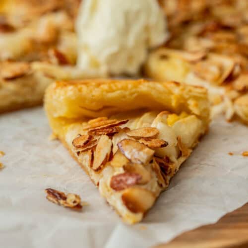 A close-up of a slice of apple galette with almond filling and a golden crust, sitting on parchment paper. A scoop of vanilla ice cream rests on top of the remaining pastry in the background.