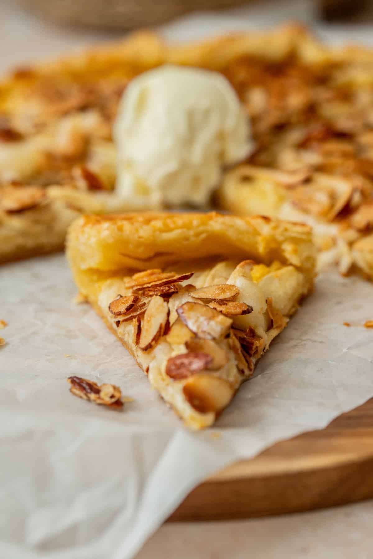 A close-up of a slice of apple galette with almond filling and a golden crust, sitting on parchment paper. A scoop of vanilla ice cream rests on top of the remaining pastry in the background.