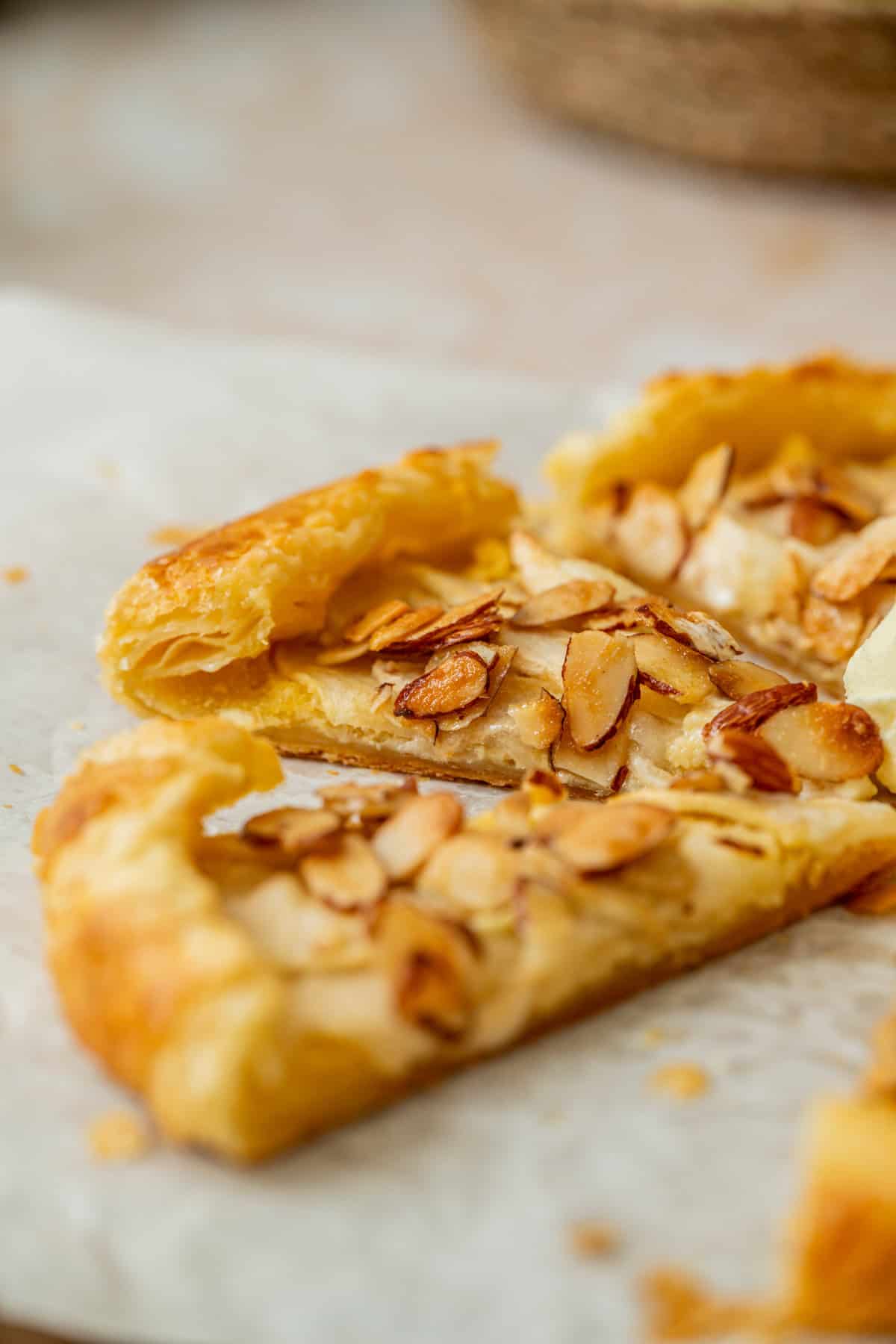 A close-up of a flaky apple galette with almond filling, topped with sliced, toasted almonds, broken into pieces and resting on parchment paper.