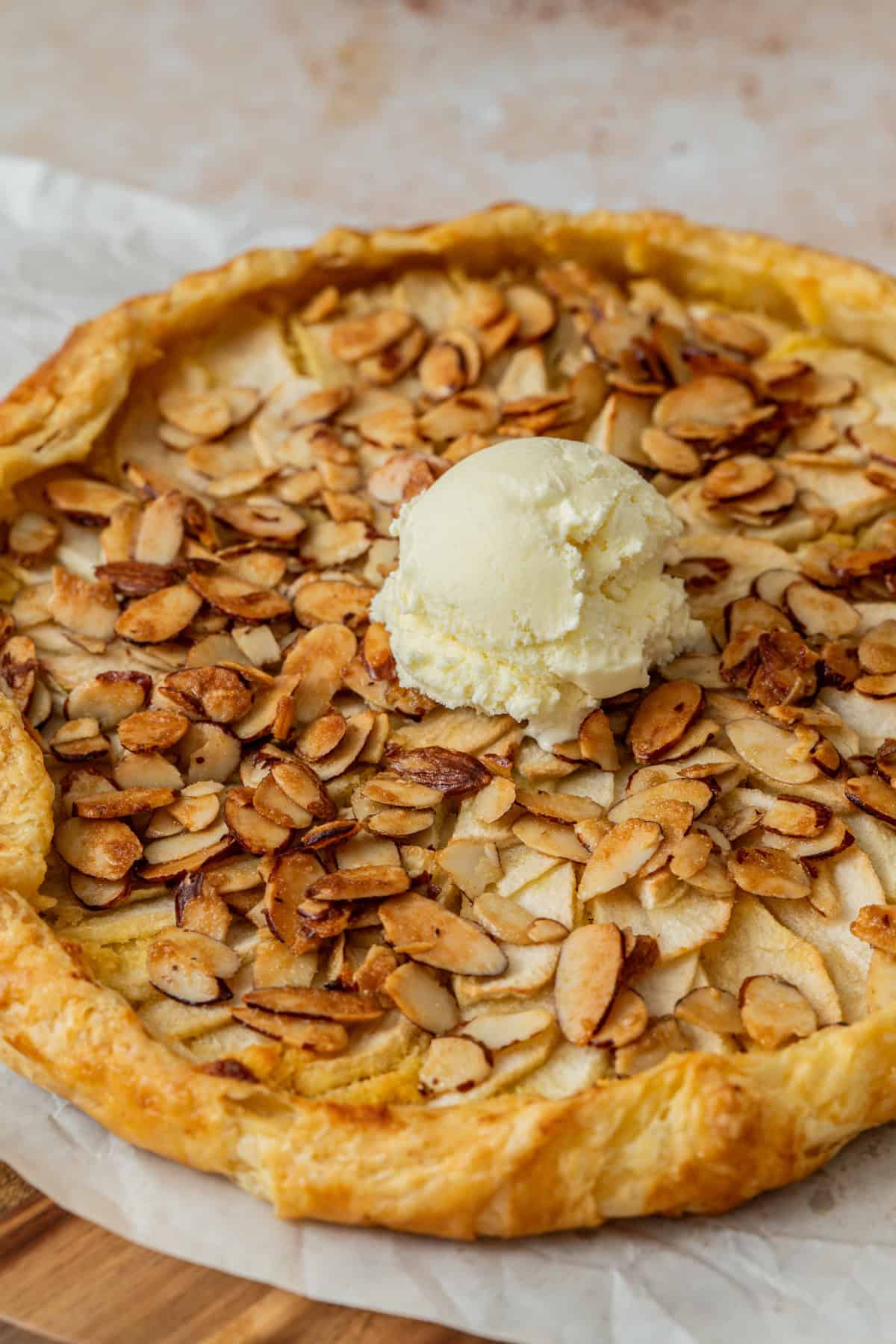 A rustic apple galette with almond filling, topped with toasted almond slices and a scoop of vanilla ice cream, sits on parchment paper, showing a flaky golden-brown crust.
