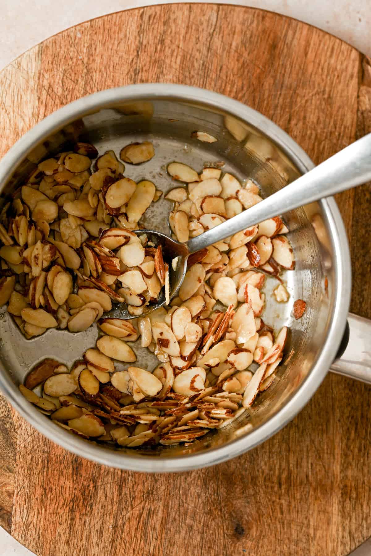 A silver saucepan containing toasted sliced almonds for an apple galette with almond filling, with a metal spoon, rests on a round wooden cutting board.