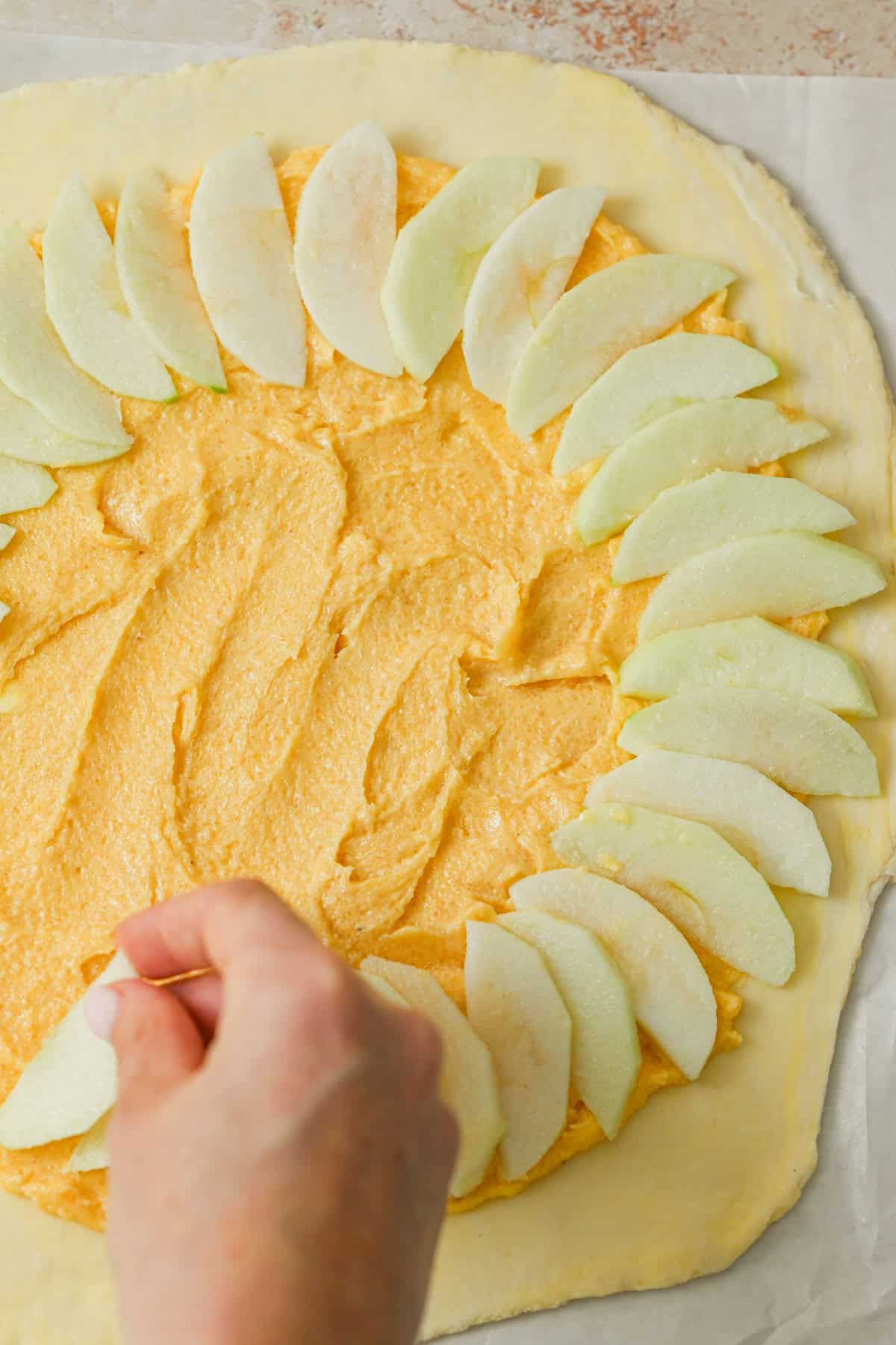 A hand arranges thin slices of green apple in a circular pattern on dough spread with almond filling, preparing an apple galette with almond filling on parchment paper.