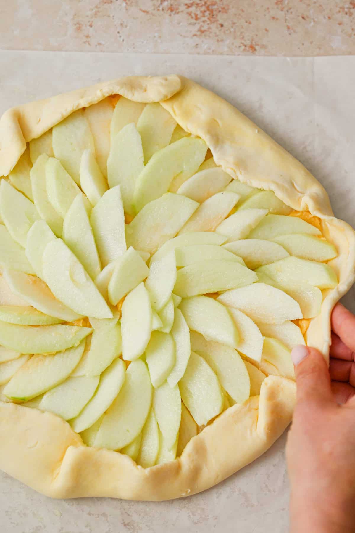 A hand folds the edge of an unbaked apple galette with almond filling, its thinly sliced green apples arranged in a circular pattern on a light countertop.