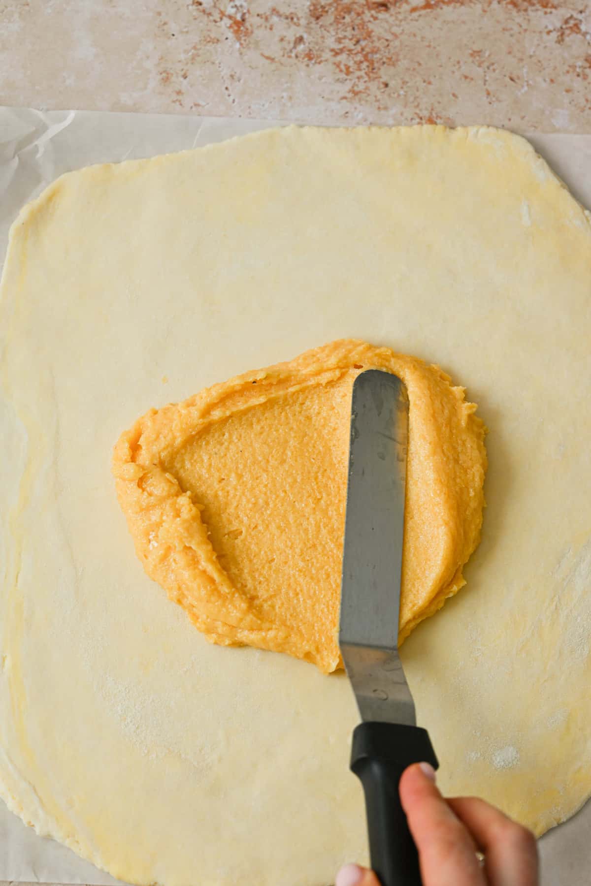 A hand spreads a thick layer of yellow almond filling onto rolled-out dough for an apple galette with almond filling, using an offset spatula on a light countertop.