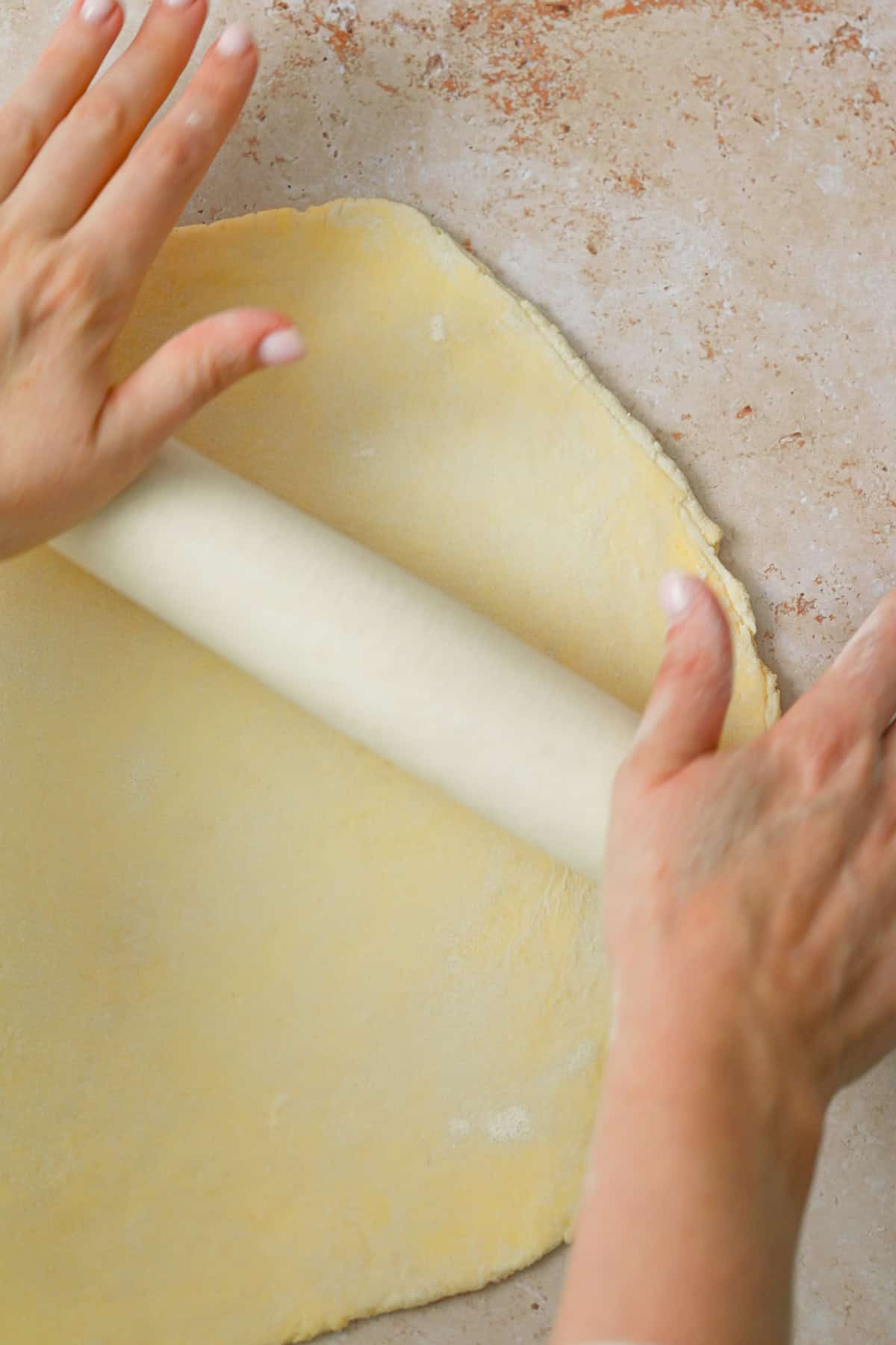 A person uses a rolling pin to flatten dough on a lightly floured surface, preparing it for baking an apple galette with almond filling.