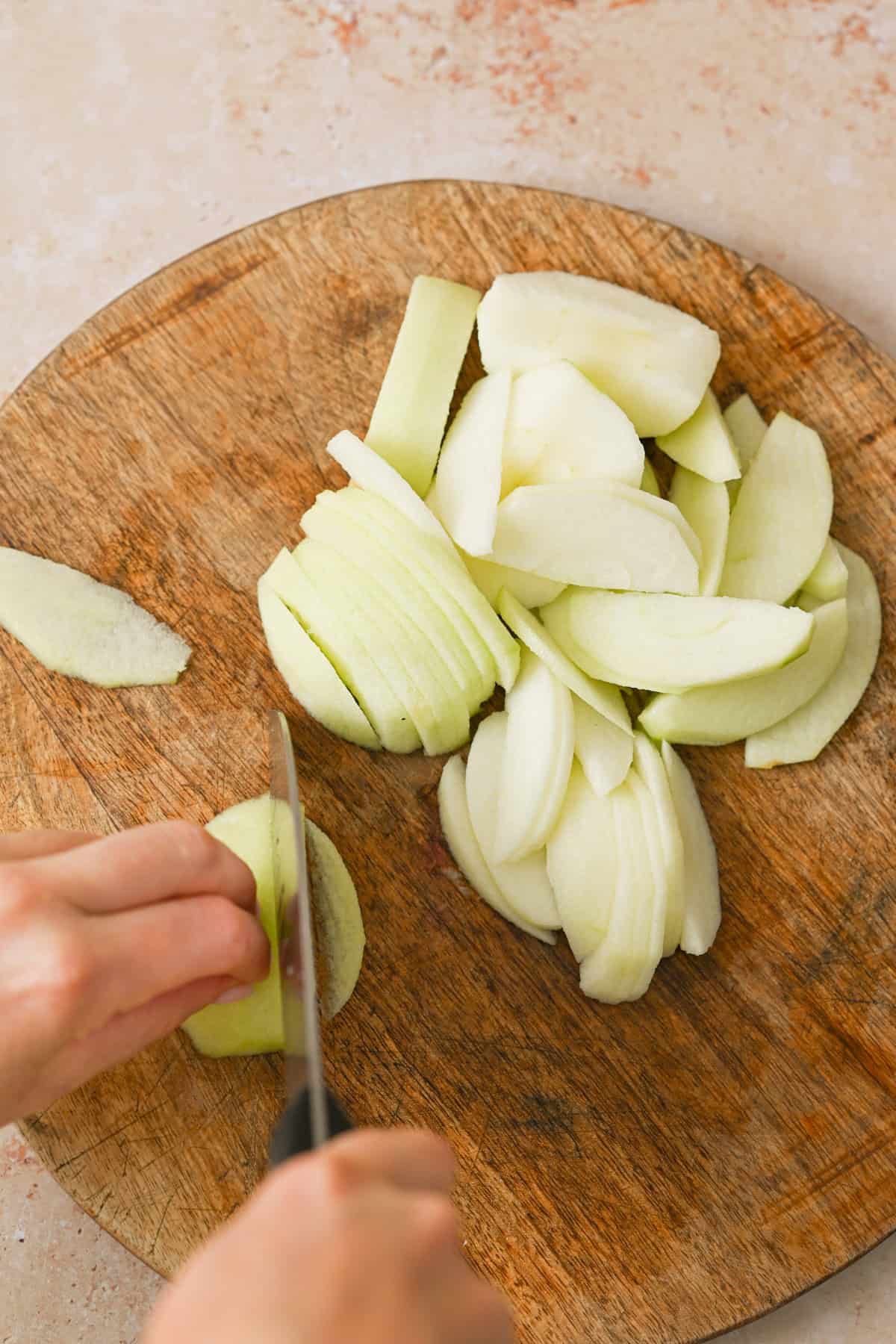 A person slices peeled green apples on a round wooden cutting board, creating a pile of thin apple slices to use in an apple galette with almond filling.