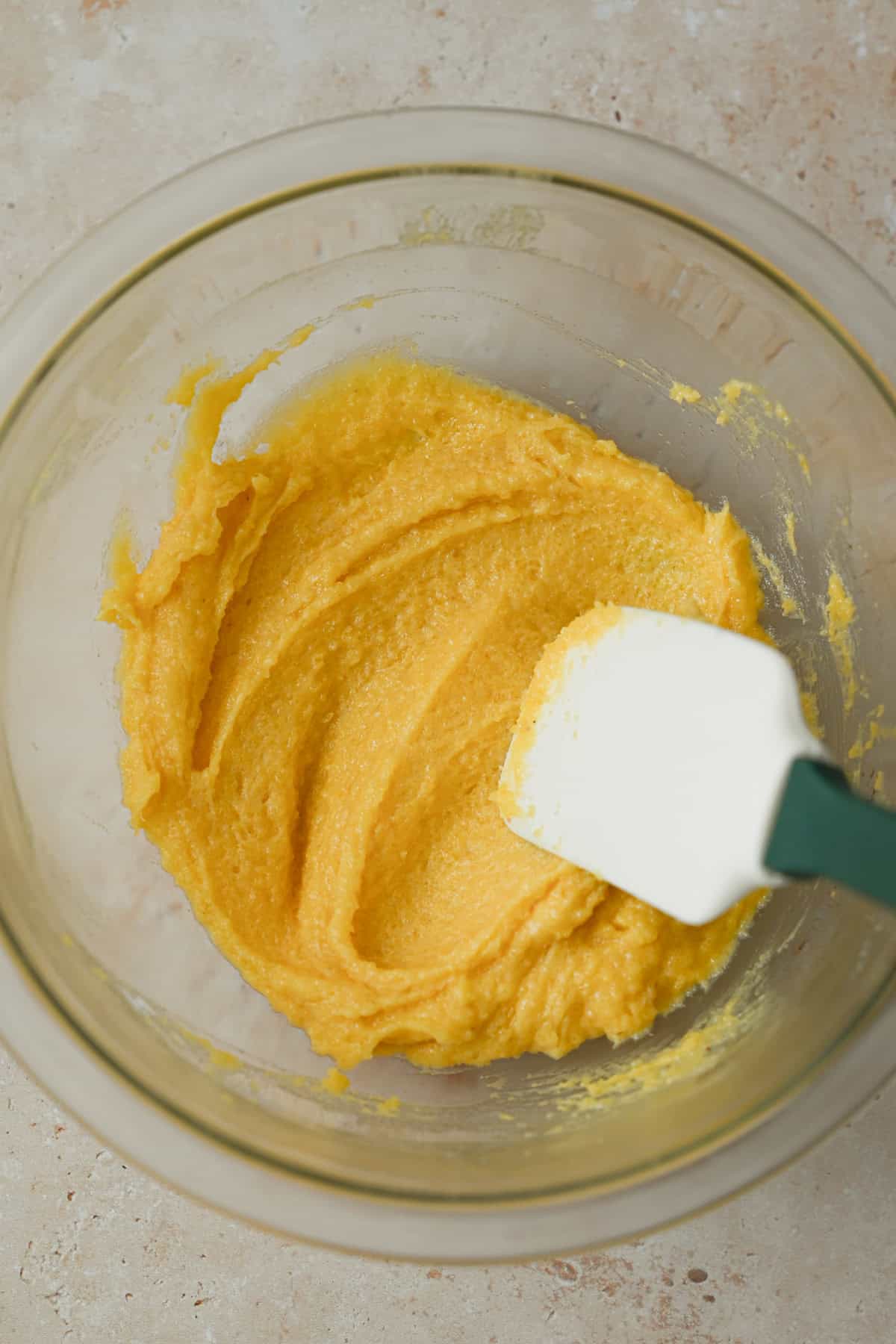 A glass bowl containing a thick, yellow batter—perhaps the base for an apple galette with almond filling—being mixed with a white and green spatula on a light countertop.