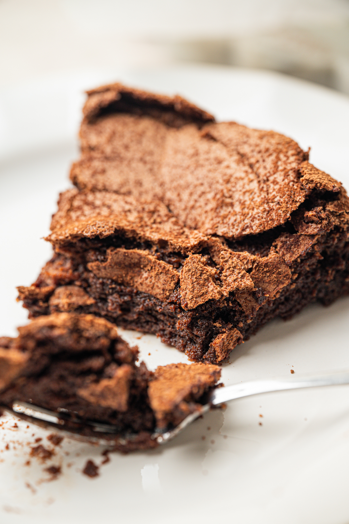 A close-up of a slice of flourless chocolate cake with a cracked, textured top on a white plate. A fork is in the foreground, breaking off a bite-sized piece of the cake.