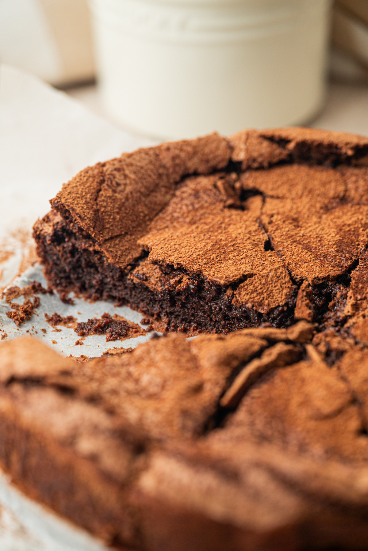 A close-up of a sliced, rustic flourless chocolate cake dusted with cocoa powder, sitting on parchment paper.
