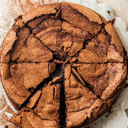 A round, cracked flourless chocolate cake dusted with cocoa powder is sliced into wedges, with one piece being lifted by a metal server.