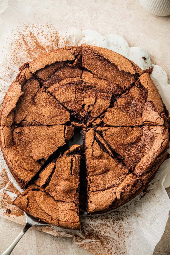 A round, cracked flourless chocolate cake dusted with cocoa powder is sliced into wedges, with one piece being lifted by a metal server.