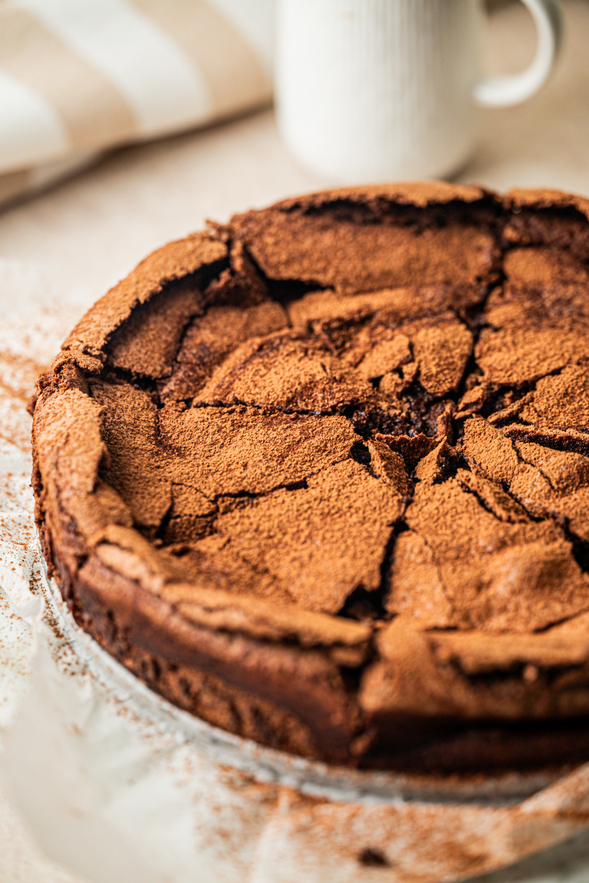 A close-up of a cracked, round flourless chocolate cake dusted with cocoa powder, sitting on parchment paper with a blurred cup and striped cloth in the background.