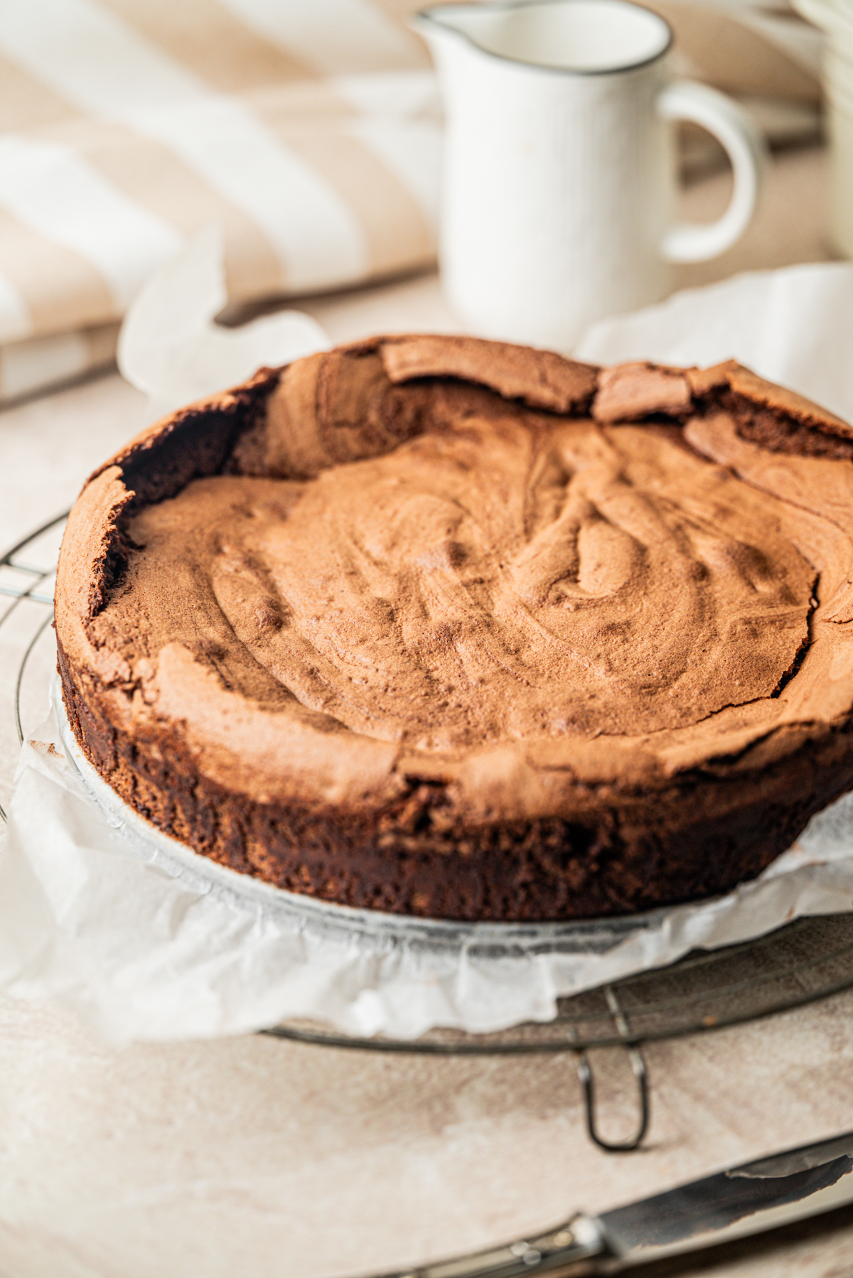 A round, homemade flourless chocolate cake with a cracked top sits on parchment paper atop a wire rack. A white pitcher and striped towel are blurred in the background.