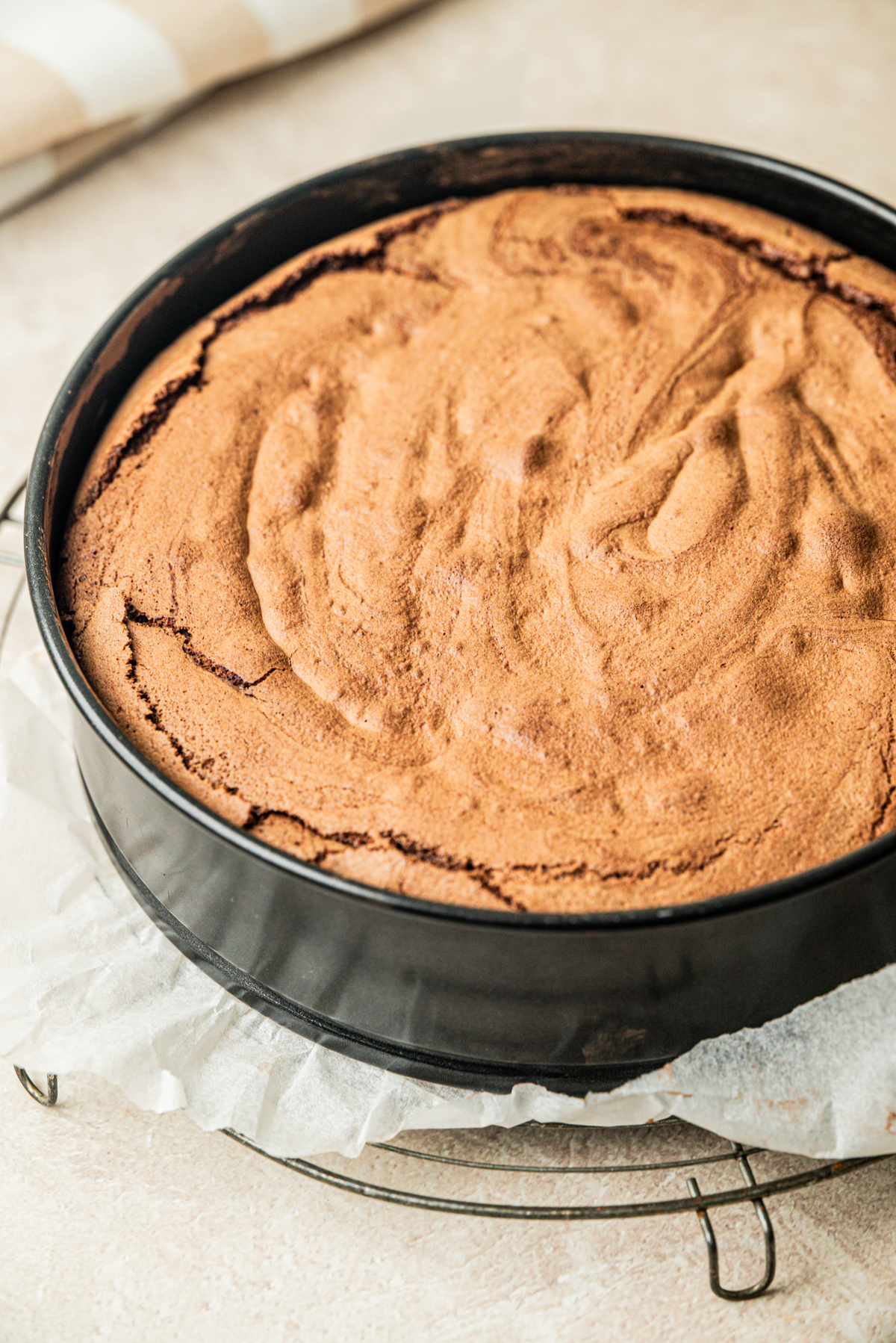 A freshly baked flourless chocolate cake in a round springform pan lined with parchment paper, resting on a metal cooling rack. The cake has a cracked, textured top.