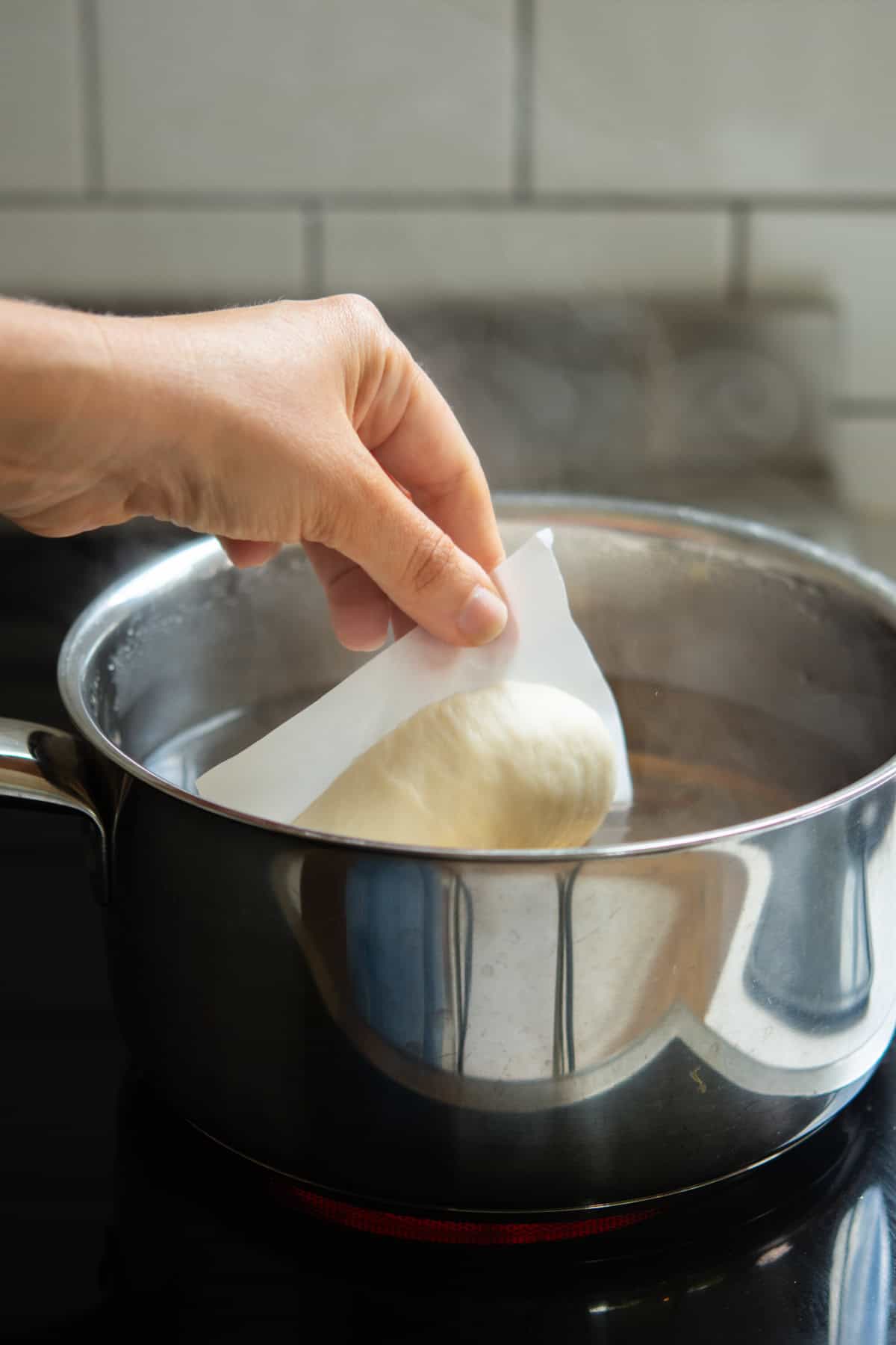 A hand lowers a piece of dough, prepared from an overnight bagel recipe, on parchment paper into a pot of boiling water on the stovetop, with steam rising and a white tiled backsplash in the background.