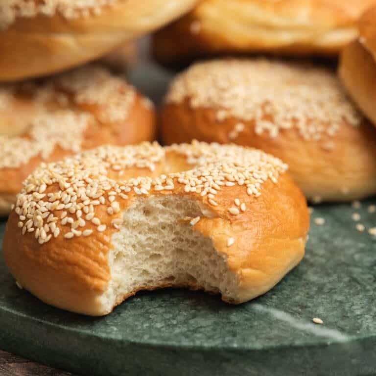 A sesame seed bagel, made from an overnight bagel recipe, sits with a bite taken out of it on a green marble surface, surrounded by more whole bagels in the background.